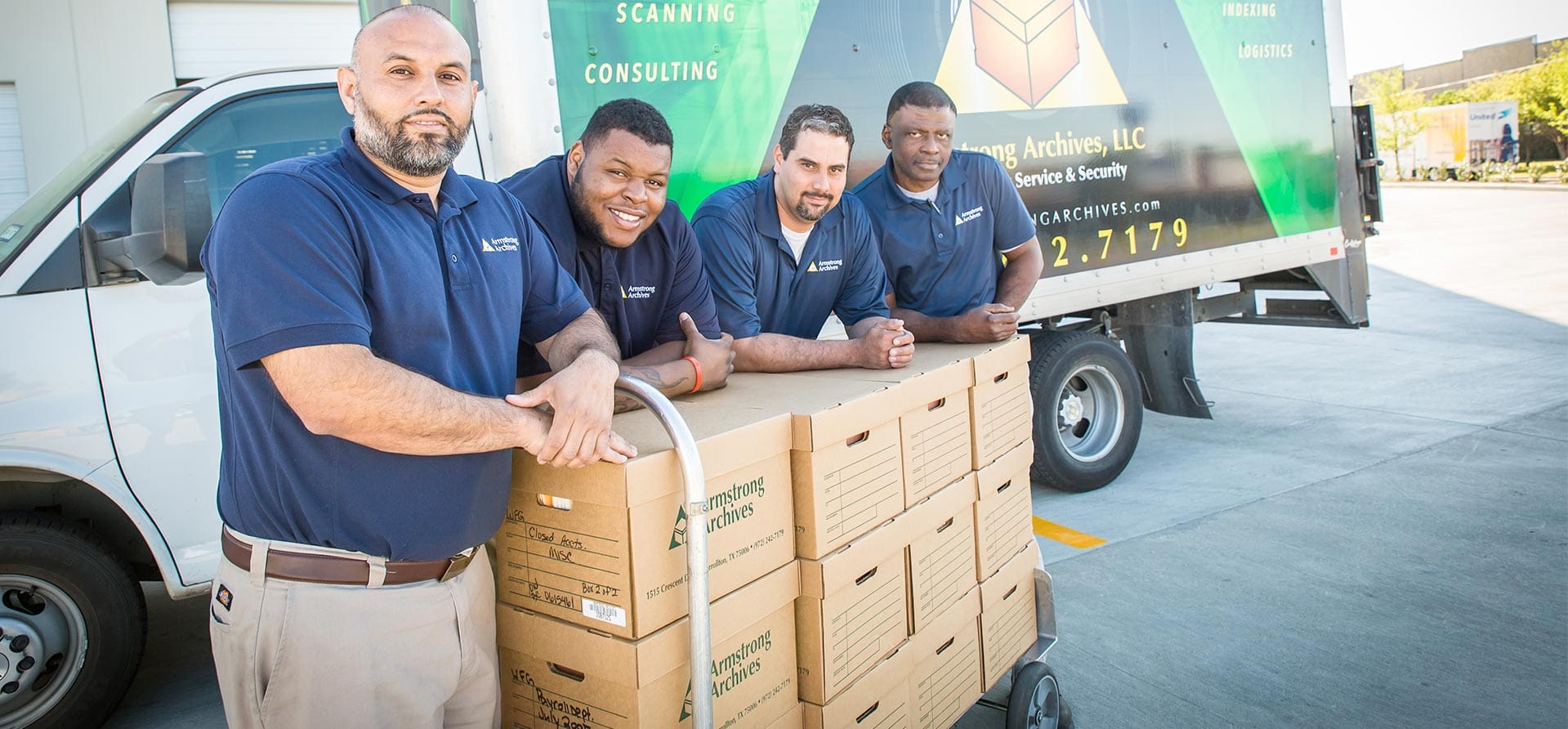 Four employees in work attire standing proudly in front of an Armstrong Archives transportation vehicle, positioned beside a stack of document storage boxes. The scene conveys teamwork, efficiency, and the secure transportation of important documents.